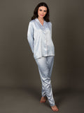 A woman standing and wearing an ice blue satin night suit with a collar shirt and an elasticated pajama. The night suit has long sleeves and consists of a shirt, pyjama pants.