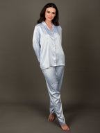 A woman standing and wearing an ice blue satin night suit with a collar shirt and an elasticated pajama. The night suit has long sleeves and consists of a shirt, pyjama pants.