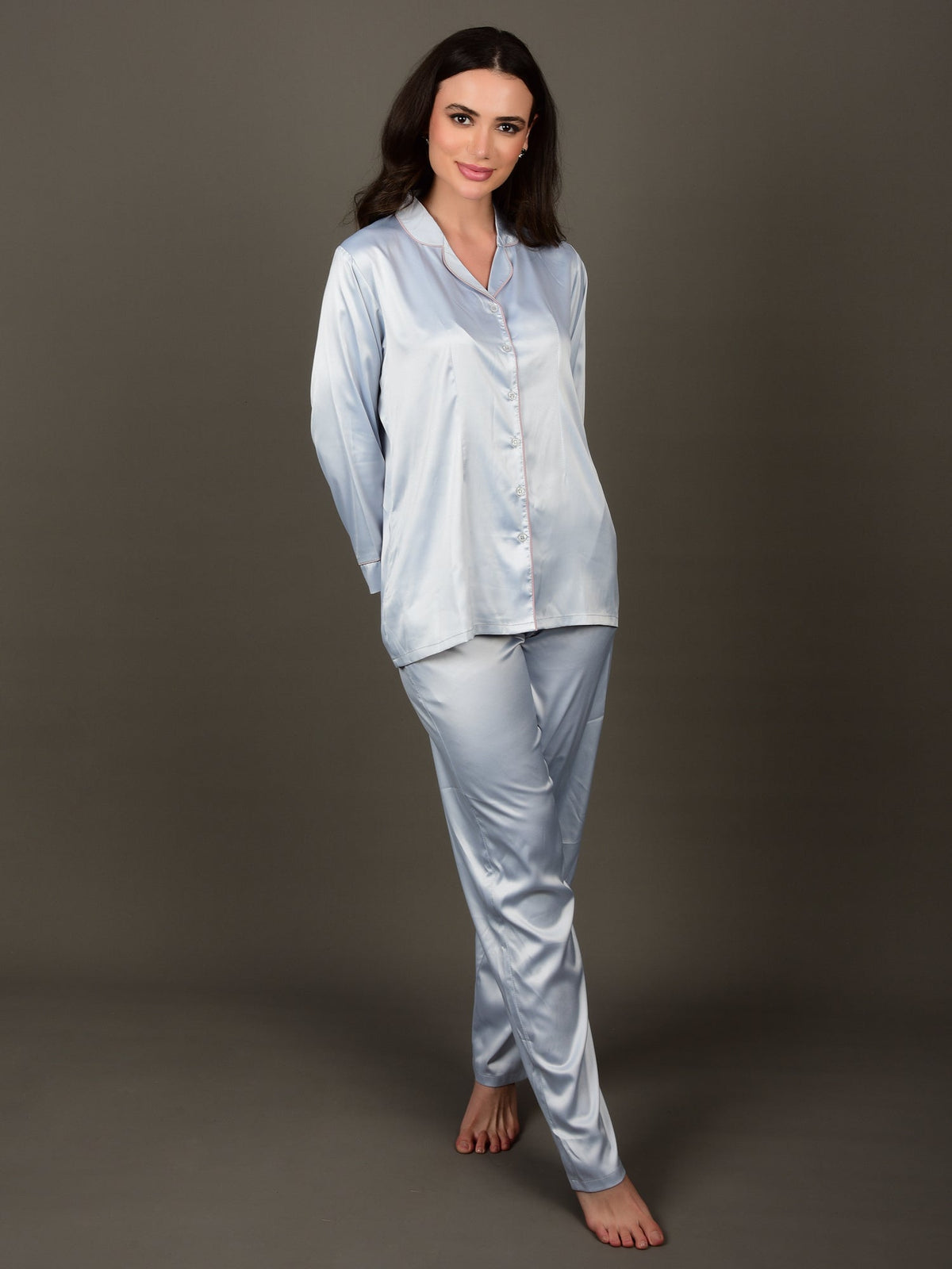 A woman standing and wearing an ice blue satin night suit with a collar shirt and an elasticated pajama. The night suit has long sleeves and consists of a shirt, pyjama pants.